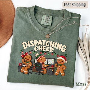 Op de afbeelding: Een mosgroen t-shirt met ronde hals en een afbeelding van drie peperkoekfiguurtjes die aan bureaus met computers en headsets werken. De tekst DISPATCHING CHEER staat boven de figuren, die versierd zijn met kerstaccessoires.