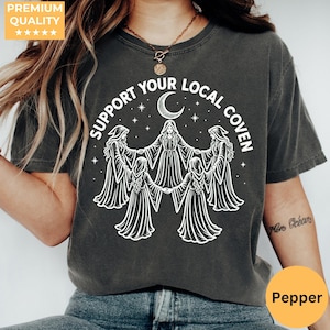 May include: A charcoal gray short-sleeve t-shirt featuring a white graphic print of four figures in hooded robes standing in a circle with hands joined. Above the figures is a crescent moon and stars with the text SUPPORT YOUR LOCAL COVEN.