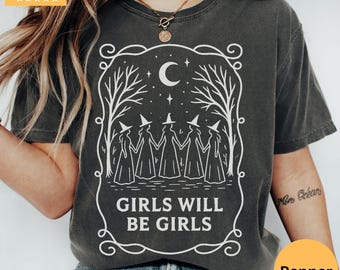 May include: Dark gray t-shirt with a white graphic of five witches holding hands under a crescent moon and stars, framed by trees. The text "GIRLS WILL BE GIRLS" is below the graphic. The shirt has a Pepper color tag.