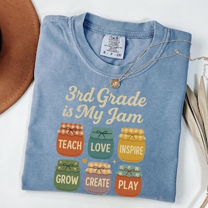 May include: A light blue t-shirt with the text "3rd Grade is My Jam" and six colorful jars with words like "Teach", "Love", "Inspire", "Grow", "Create", and "Play".