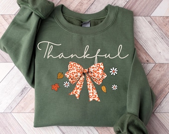 Könnte beinhalten: Ein olivgrünes Sweatshirt mit dem Wort "Thankful" in weißer Schrift. Das Design umfasst eine Schleife mit Leopardenmuster, Herbstblätter und kleine weiße Gänseblümchen. Ein gemütliches Kleidungsstück zum Thema Herbst.