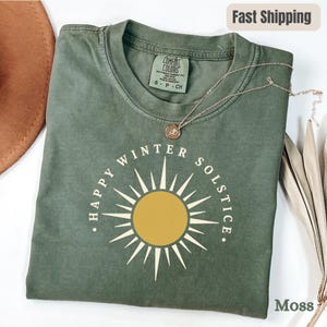 以下が含まれることがあります： モスグリーンのTシャツで、放射状の白い線を持つ黄色い太陽のグラフィックと、円状に配置された「HAPPY WINTER SOLSTICE」の文字がプリントされています。クルーネックで、襟の内側にはComfort Colorsのブランドラベルが付いています。