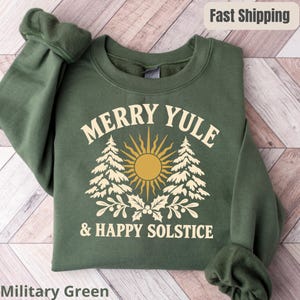 Może przedstawiać: Wojskowa zielona bluza z okrągłym dekoltem i kremowym nadrukiem graficznym. Wzór zawiera napis MERRY YULE & HAPPY SOLSTICE, centralną ikonę słońca, dwie sosny oraz liście ostrokrzewu. Bluza posiada długie rękawy oraz ściągacz przy dekolcie.