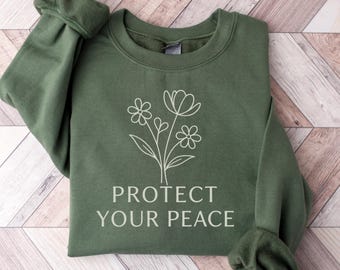 May include: An olive green sweatshirt with the words "PROTECT YOUR PEACE" in white block letters. A white floral design is above the text. The sweatshirt is folded on a light brown wooden surface.