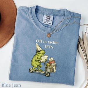 May include: Blue jean Comfort Colors t-shirt with the text "Off to tackle IEPs." The shirt features a whimsical illustration of a green frog wearing a wizard hat and riding a scooter, carrying a stack of books and an IEP form.