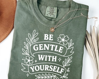 Może przedstawiać: Koszulka Comfort Colors w kolorze mchu z białym napisem "BE GENTLE WITH YOURSELF" i kwiatowymi akcentami. Złoty naszyjnik z wisiorkiem jest ułożony na koszulce. Brązowy kapelusz i suszone liście znajdują się w tle.