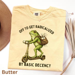 May include: Pale yellow t-shirt featuring a green frog on a scooter, with a backpack. Text reads "OFF TO GET RADICALIZED BY BASIC DECENCY." The shirt has a Comfort Colors label. A necklace and dried plant are also visible.