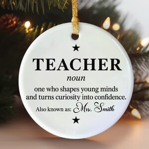 以下が含まれることがあります： 黒い文字で「TEACHER noun one who shapes young minds and turns curiosity into confidence. Also known as: Mrs. Smith.」と書かれた白い円形のセラミック製オーナメント。2つの黒い星と吊り下げ用の金色のリボンが付いています。