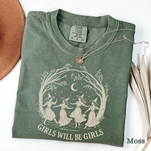 May include: A moss green t-shirt with a round neck and short sleeves. The shirt features a graphic of four women in witch hats dancing under a crescent moon, surrounded by a tree branch design. The text "GIRLS WILL BE GIRLS" is printed below the graphic. A gold necklace is draped across the shirt.