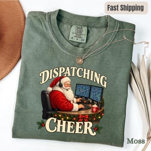May include: A moss green t-shirt featuring a graphic of Santa Claus wearing a headset and sitting at a desk with two computer monitors. The text "DISPATCHING CHEER" is printed in a cream-colored font above and below the graphic of Santa.
