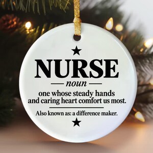 Może przedstawiać: Okrągła biała ceramiczna ozdoba z czarnym napisem: NURSE noun one whose steady hands and caring heart comfort us most. Also known as: a difference maker. Ozdoba zawiera dwie czarne gwiazdki oraz złotą wstążkę do zawieszenia.
