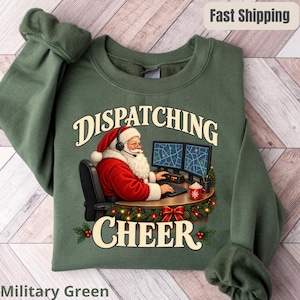 Könnte beinhalten: Ein militärgrüner Rundhals-Pullover mit einer Grafik vom Weihnachtsmann, der ein Headset trägt und an einem Schreibtisch mit zwei Computermonitoren sitzt. Der Text "DISPATCHING CHEER" ist in cremefarbener Schrift über und unter dem Bild gedruckt.