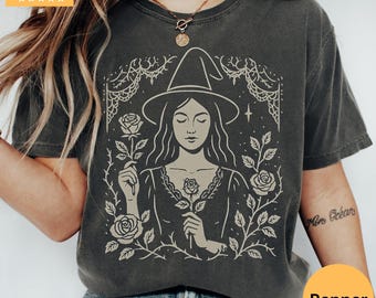 May include: Dark gray t-shirt featuring a beige illustration of a woman wearing a witch hat and holding a rose. The design is surrounded by roses and vines. The shirt has a Pepper color label.
