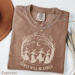 May include: A brown short-sleeve t-shirt with a crew neckline and a Comfort Colors label. The graphic features four figures in witch hats dancing under a crescent moon and tree branches, with the text "GIRLS WILL BE GIRLS" printed below.