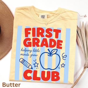 Op de afbeelding: Een botergeel t-shirt met een lichtblauwe verticaal gestreepte achtergrond. De afbeelding bevat de rode tekst "FIRST GRADE CLUB" en de blauwe tekst "helping little minds grow" naast een blauw appelicoon, een potlood en sterren.