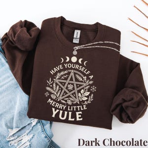 以下が含まれることがあります： ダークチョコレート色のスウェットシャツ。白いグラフィックには五芒星、三日月、そして「Have Yourself a Merry Little Yule」の文字が描かれています。シルバーのネックレスがスウェットシャツの上にあります。「Dark Chocolate」の文字が下にあります。