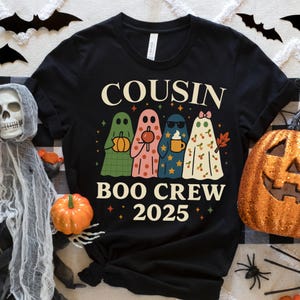 May include: Black t-shirt featuring the text "COUSIN BOO CREW 2025" and a graphic of four ghost figures. The ghosts are in green, pink, blue, and white, each holding a different item. Halloween-themed clothing.