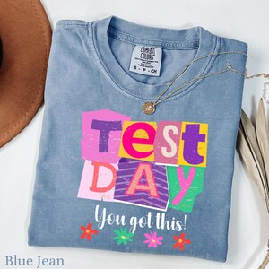 Könnte beinhalten: Ein blaues Jeans-T-Shirt von Comfort Colors mit den Worten "Test Day" in bunten Blockbuchstaben. Darunter steht "You got this!" mit floralen Akzenten. Eine goldene Halskette und ein brauner Hut sind ebenfalls zu sehen.