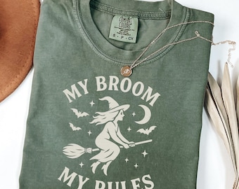 May include: A moss green t-shirt with the phrase "MY BROOM MY RULES" in white, featuring a witch on a broom, bats, stars, and a crescent moon. A gold necklace with a pendant rests on the shirt.