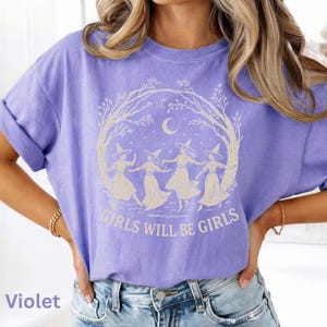 May include: A violet short-sleeve t-shirt featuring a cream-coloured graphic of four figures wearing pointed hats dancing in a circle under a crescent moon and tree branches, with the text "GIRLS WILL BE GIRLS" printed in a bold font below the design.