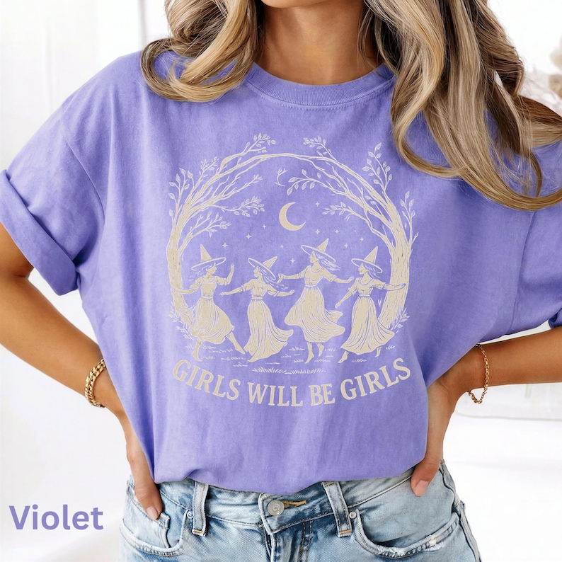 May include: A violet short-sleeve t-shirt featuring a cream-coloured graphic of four figures wearing pointed hats dancing in a circle under a crescent moon and tree branches, with the text "GIRLS WILL BE GIRLS" printed in a bold font below the design.