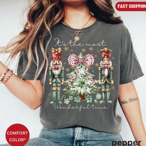May include: A gray t-shirt with a Christmas-themed graphic. The design features two nutcrackers, a Christmas tree, and the text "It's the most Wonderful time". The tree has a large red and white polka dot bow.