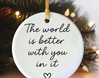 May include: A white ceramic Christmas ornament with the message "The world is better with you in it" in black script, and a heart symbol. It hangs from a gold ribbon, with a blurred background of Christmas lights and greenery.