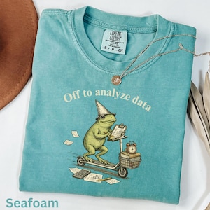 Puede incluir: Camiseta verde mar con la frase "Off to analyze data" y un gráfico de una rana en un scooter. La rana lleva un sombrero de mago y sostiene una carpeta. También se ven un collar y un sombrero marrón.