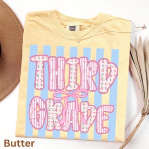 Pode incluir: Uma camiseta amarela Comfort Colors com as palavras "THIRD GRADE" em um design rosa e branco com cerejas. A camisa tem listras verticais azuis e um laço rosa. A palavra "Butter" está na parte inferior.