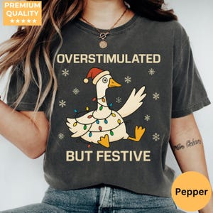 Peut inclure: T-shirt gris foncé avec un dessin d'oie de dessin animé portant un bonnet de Père Noël et des guirlandes lumineuses. Le texte sur le t-shirt se lit "OVERSTIMULATED BUT FESTIVE" avec des flocons de neige. Le t-shirt a un aspect vintage et usé.