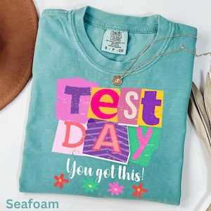 Könnte beinhalten: Türkisgrünes T-Shirt mit den Worten "Test Day" in bunten Blockbuchstaben und dem Satz "You got this!" in Weiß. Das Shirt hat einen Rundhalsausschnitt und kurze Ärmel. Eine goldene Halskette und Blumen sind ebenfalls zu sehen.