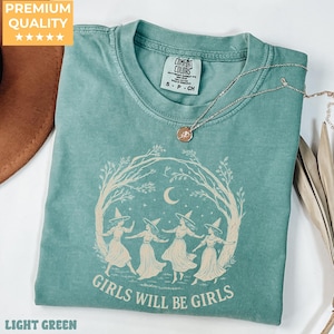 May include: A sage green short-sleeve t-shirt featuring a cream-coloured graphic of four figures in witch hats dancing under a crescent moon and tree branches. The text "GIRLS WILL BE GIRLS" is printed below the graphic. A gold-tone necklace rests on top.