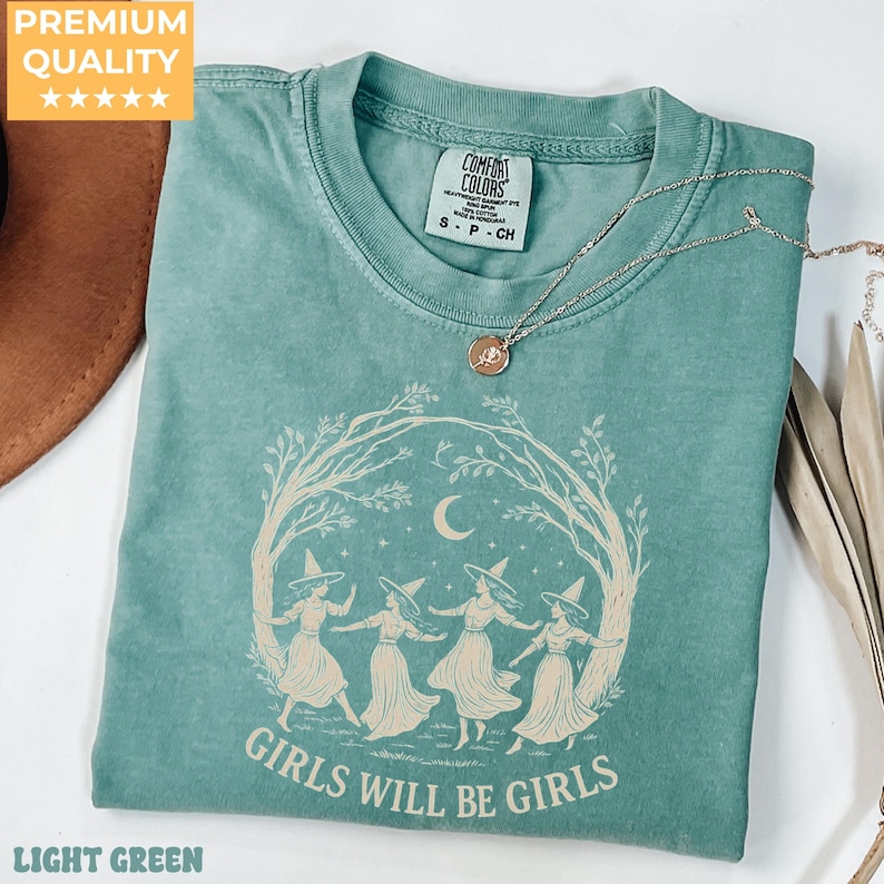 May include: A sage green short-sleeve t-shirt featuring a cream-coloured graphic of four figures in witch hats dancing under a crescent moon and tree branches. The text "GIRLS WILL BE GIRLS" is printed below the graphic. A gold-tone necklace rests on top.