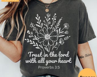 Peut inclure: T-shirt gris foncé avec un motif floral blanc et le texte "Trust in the lord with all your heart" de Proverbes 3:5. Le t-shirt a un col rond et des manches courtes. Le t-shirt est de couleur Pepper.