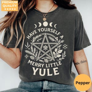 以下が含まれることがあります： 「HAVE YOURSELF A MERRY LITTLE YULE」の白文字がプリントされた濃いグレーのTシャツ。デザインには五芒星、三日月、植物の要素が含まれています。Tシャツはクルーネックで半袖です。