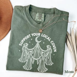 May include: An olive green t-shirt featuring a white graphic of five figures in robes holding hands in a circle under a crescent moon and stars. The text reads "SUPPORT YOUR LOCAL COVEN". The shirt has a Comfort Colors label at the neck.
