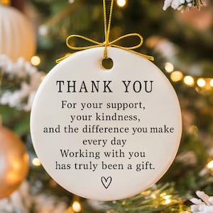 Könnte beinhalten: Ein weißer, runder Ornament mit dem schwarzen Schriftzug "THANK YOU". Das Ornament enthält eine Dankesbotschaft und ein Herzsymbol. Es wird von einem goldenen Faden gehalten und vor einem verschwommenen Weihnachtsbaumhintergrund präsentiert.