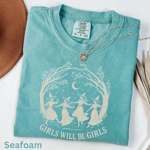 May include: A seafoam green short-sleeve t-shirt featuring a cream-coloured graphic of four figures in pointed hats dancing in a circle under a crescent moon and stars, framed by tree branches, with the text GIRLS WILL BE GIRLS printed below.