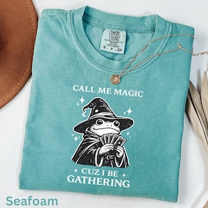May include: Seafoam green t-shirt with the text "CALL ME MAGIC" and "CUZ I BE GATHERING". The shirt features a cartoon frog wearing a wizard hat and cape, holding cards. A gold necklace is draped across the shirt.