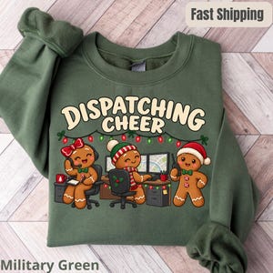 Op de afbeelding: Een militair groen sweatshirt met een afbeelding van drie peperkoekmannetjes die in een kantoor werken met de tekst DISPATCHING CHEER. Het ontwerp bevat kerstverlichting, computermonitoren en een radio in feestelijke kleuren.