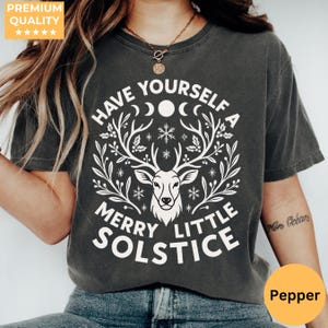 以下が含まれることがあります： 白いグラフィックが特徴の濃い灰色のTシャツ。鹿の頭、角、そして「HAVE YOURSELF A MERRY LITTLE SOLSTICE」というテキストが描かれています。 シャツはクルーネックと半袖です。 「Pepper」という単語が入った黄色の円が右下にあります。