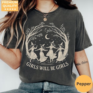 May include: A charcoal grey short-sleeve t-shirt featuring a cream-coloured graphic of four figures in pointed hats dancing in a circle under a crescent moon and tree branches, with the text "GIRLS WILL BE GIRLS" printed in a bold serif font below.