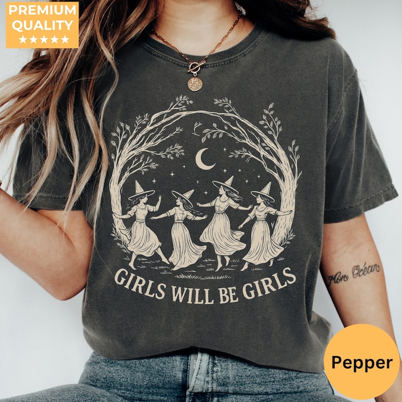 May include: A charcoal grey short-sleeve t-shirt featuring a cream-coloured graphic of four figures in pointed hats dancing in a circle under a crescent moon and tree branches, with the text "GIRLS WILL BE GIRLS" printed in a bold serif font below.