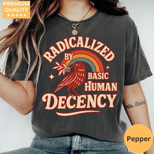Könnte beinhalten: Dunkelgraues T-Shirt mit dem Text "RADICALIZED BY BASIC HUMAN DECENCY" in Retro-Schrift. Ein roter Vogel mit einem Zweig ist unter einem Regenbogen zentriert. Das Shirt hat einen Vintage-Look.