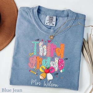 May include: A blue jean-colored t-shirt with the words "Third Grade" in colorful, patterned letters. The shirt features school-themed graphics like pencils, paperclips, and a bow. The name "Mrs. Wilson" is printed below the graphic. A gold necklace is draped across the shirt.