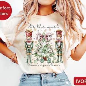 Puede incluir: Camiseta color marfil con un diseño navideño. El diseño presenta dos figuras de cascanueces flanqueando un árbol de Navidad con un gran lazo. El texto "It's the most wonderful time" está impreso en la camiseta.