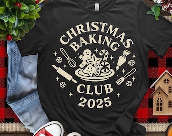 Könnte beinhalten: Schwarzes T-Shirt mit Rundhalsausschnitt und cremefarbenem Aufdruck "CHRISTMAS BAKING CLUB 2025". Das Design zeigt Backutensilien und Lebkuchen. Das Shirt wird auf einer Holzoberfläche mit festlicher Dekoration präsentiert.