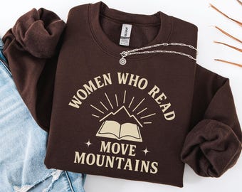 May include: A dark chocolate-colored sweatshirt with the text "WOMEN WHO READ MOVE MOUNTAINS" in a cream color. The design includes an open book, mountains, and radiating lines. A silver necklace is draped across the sweatshirt.