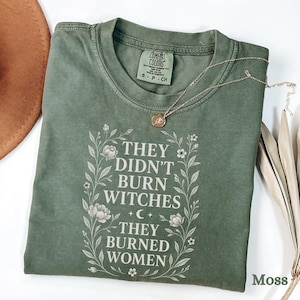 Puede incluir: Una camiseta verde musgo con el texto blanco "THEY DIDN'T BURN WITCHES THEY BURNED WOMEN", enmarcado por ilustraciones florales. Un collar dorado con un colgante descansa sobre la camiseta. La camiseta está etiquetada como "Comfort Colors".