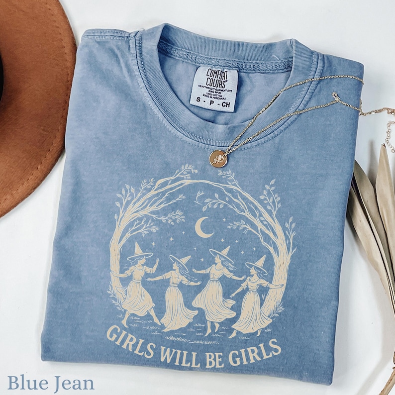 May include: A blue jean coloured t-shirt featuring a cream-coloured graphic of four figures in pointed hats dancing in a circle under a crescent moon and tree branches. The text "GIRLS WILL BE GIRLS" is printed below the graphic. A tag reads Comfort Colors.